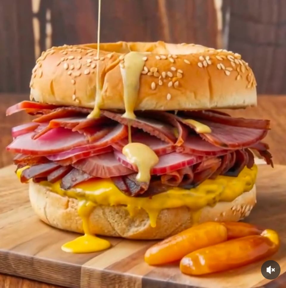 An AI image of a pastrami bagel with two awful carrot-like glazy things next to it. It's covered in two kinds of weird yellow sauce and it looks just wrong enough that it's not appetizing and it looks kinda nightmarish.